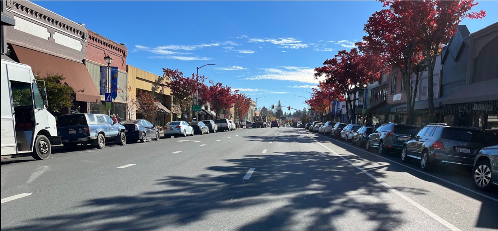 Main Street in downtown Sebastopol today, looking along the corridor: a wide roadway with parked cars on both sides, narrow sidewalks, and street trees in autumn colour.